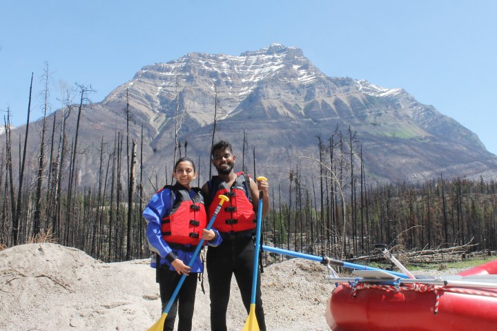 Two people in life jackets holding paddles with mountain backdrop and raft nearby.