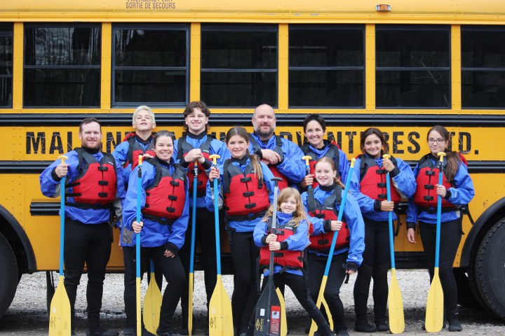 Group in red life jackets holding paddles in front of yellow school bus.