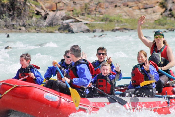 Group of people in red raft wearing life jackets, smiling and waving while white-water rafting.