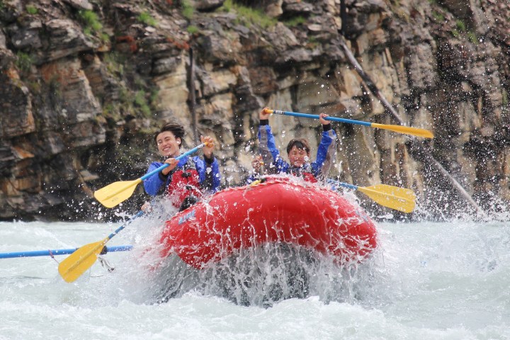 Two people rafting in a red inflatable boat on a rapid river, smiling and holding paddles.