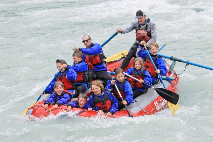 Group of people in a red raft paddling on a river with smiles and life jackets.