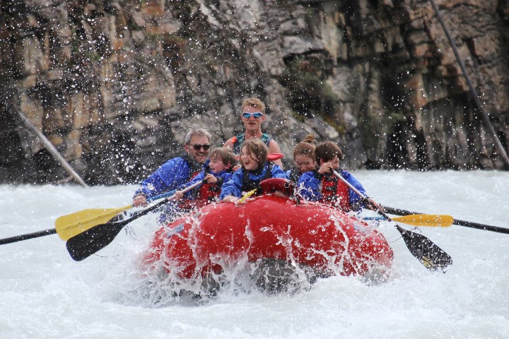 Group of people white-water rafting on a river in a red raft, with rocky cliffs in the background.