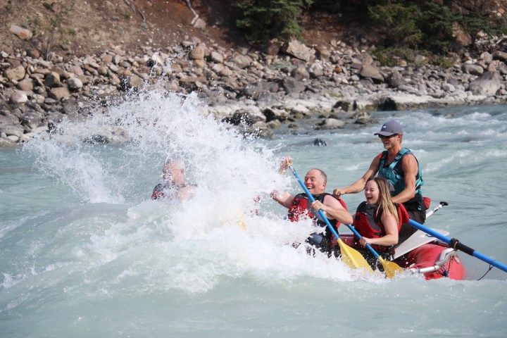 Four people whitewater rafting, splashed by water, wearing life vests and helmets on a rocky river.