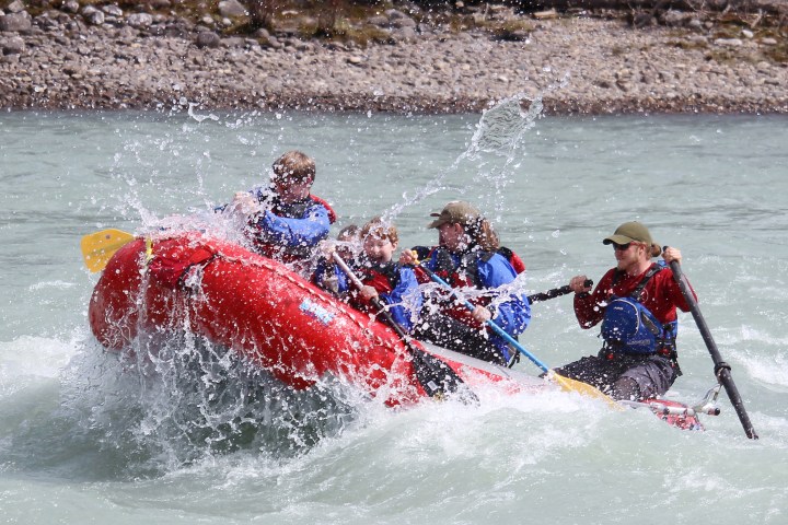 Group of people whitewater rafting in a red inflatable boat, splashing through water.