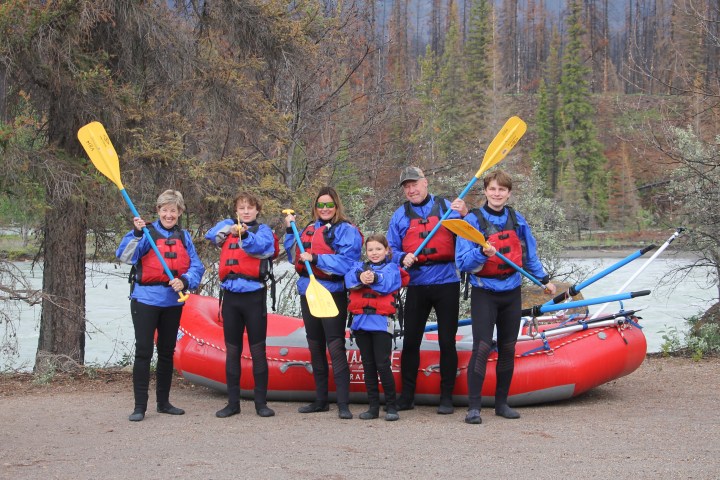 Group of six people in rafting gear posing with paddles in front of a red raft and forested river backdrop.
