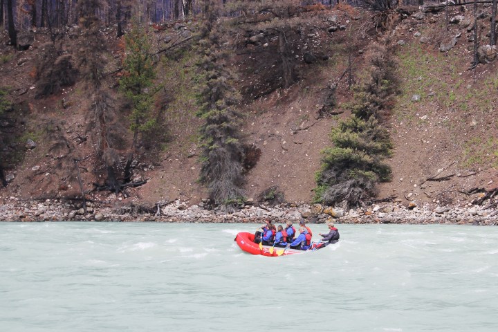 Group whitewater rafting in a red raft on a river beside a rocky forested bank.