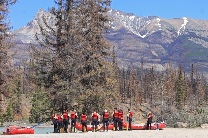 Group in red life jackets beside rafts near trees with mountains in the background.