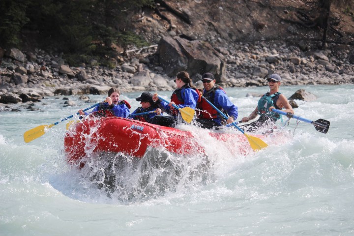 Group of people whitewater rafting in a red raft on a rocky river.