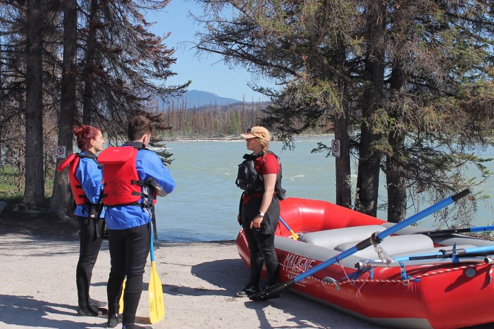 Three people in rafting gear standing by a red raft near a river surrounded by trees.