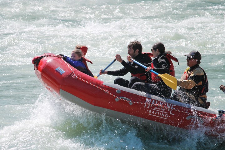 Four people white-water rafting in red inflatable boat on rough river.
