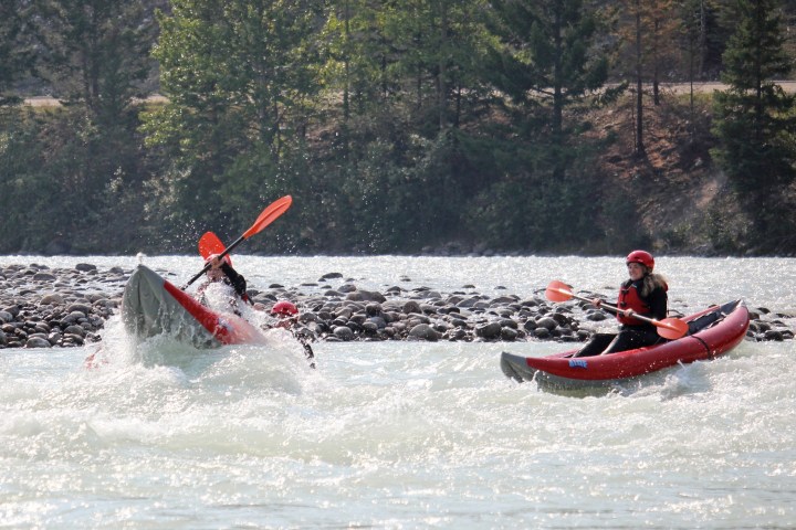 Two people kayaking on a river, one kayak splashing through whitewater, surrounded by trees.