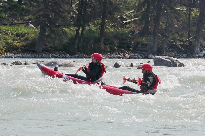 Two people in helmets kayaking on a rough river with trees in the background.