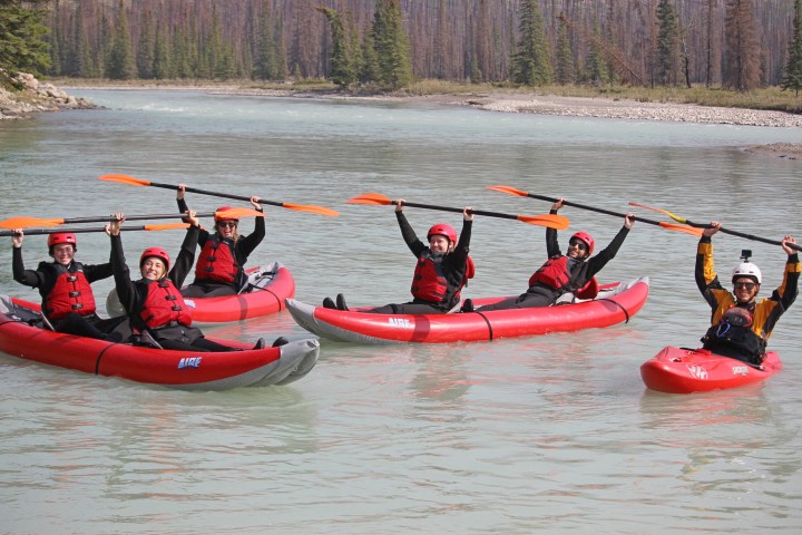 Five kayakers in red gear holding paddles, smiling on a calm river with pine trees in the background.