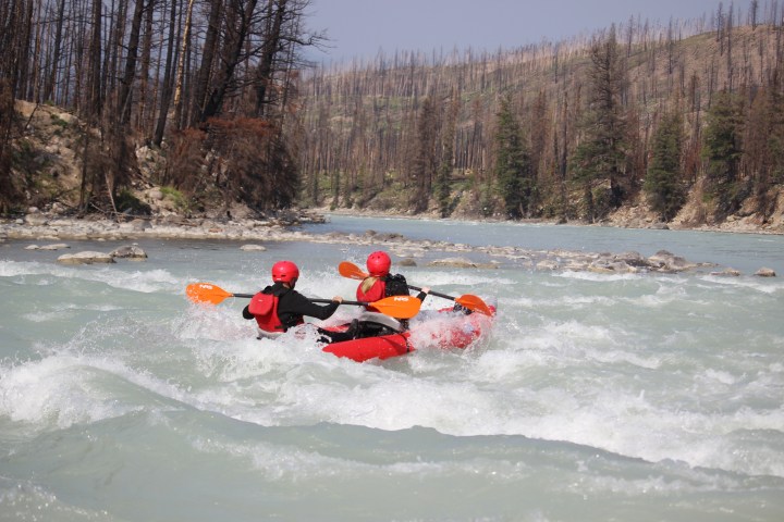 Two kayakers in red gear navigate rapids on a wide river surrounded by forested hills.