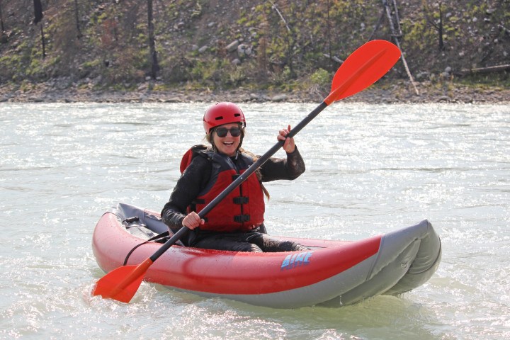 Person kayaking on a river, wearing a red helmet and life jacket, holding a paddle.