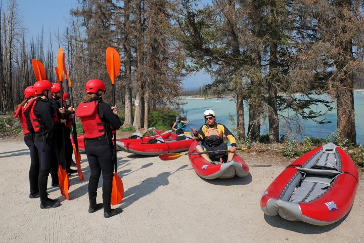 Group in red gear learns kayaking beside inflatable kayaks and a river, trees in the background.