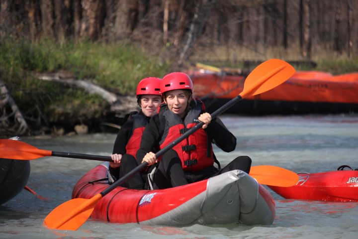 Two people kayaking on a river wearing red helmets and life vests.