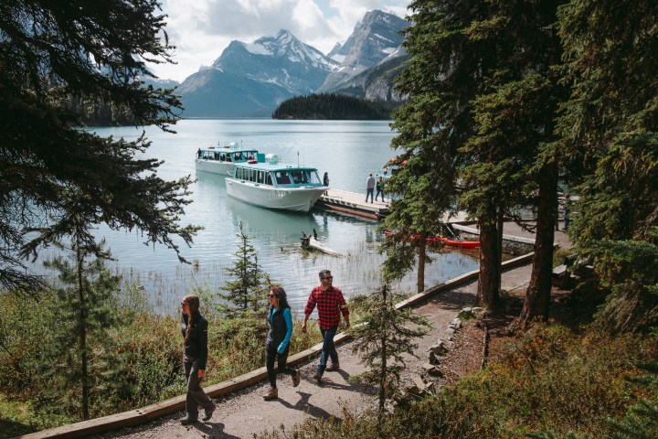 People walking by a lake with a boat docked and mountains in the background.