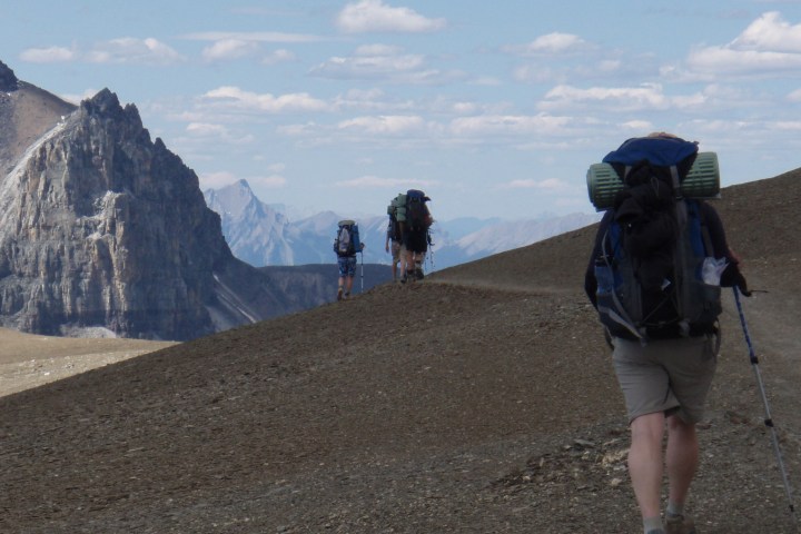 Three hikers with backpacks walking on a mountain trail under a blue sky with clouds.