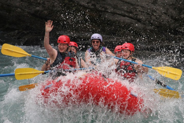 Group of people white-water rafting in a red raft, wearing helmets and life jackets.