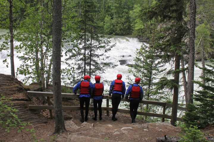 Four people in helmets and life jackets overlook a river with rapids.