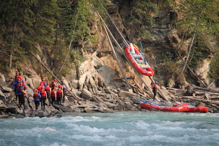 Group of rafters in helmets near river with rafts on rocky shore and trees.