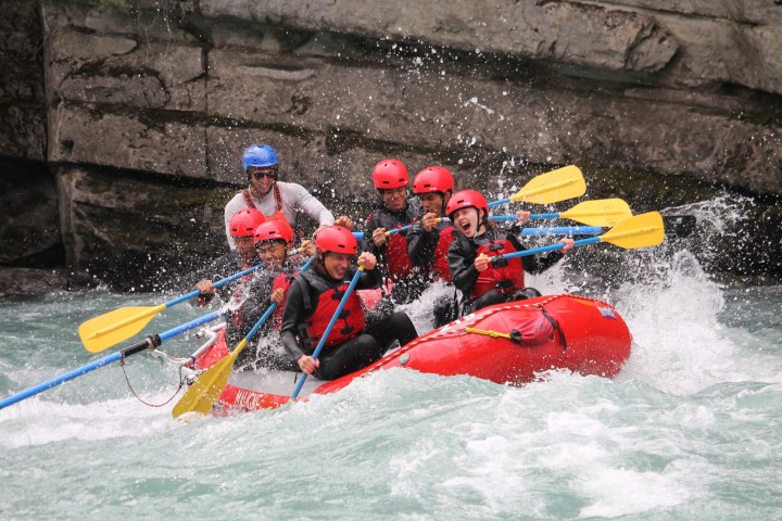 Group of people whitewater rafting on turbulent river in a red raft, wearing helmets and life jackets.