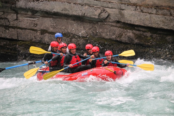 Group of people whitewater rafting in a red raft, wearing helmets and life jackets.