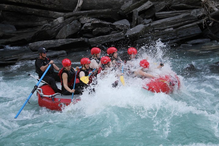 Group of people whitewater rafting on a river, wearing helmets and life jackets, splashing through waves.