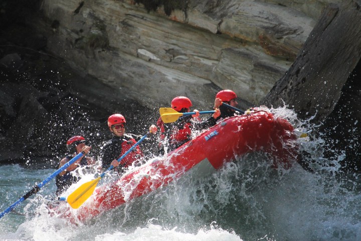Group white-water rafting in a red raft, wearing helmets and life jackets, splashing through rapids.