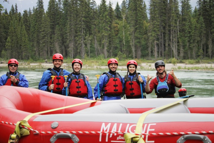 Six people in helmets and vests stand beside raft in front of river and trees.