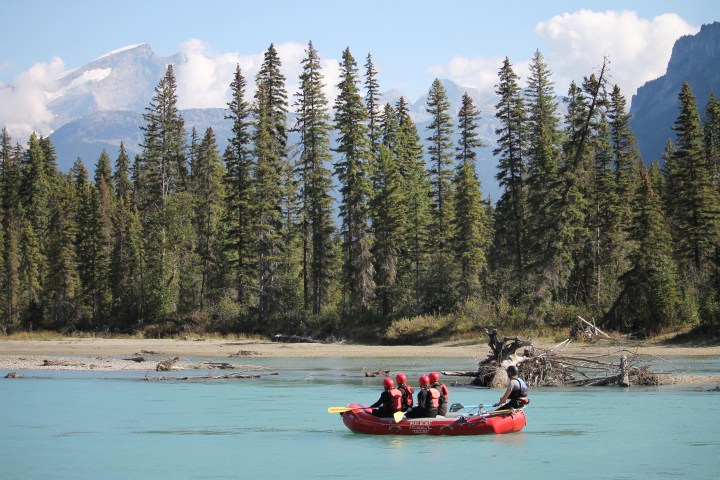 People in a red raft paddling on a turquoise river with trees and mountains in the background.