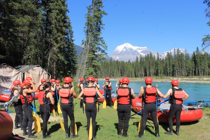 Group of people with helmets and life jackets preparing for rafting by a river with mountain view.