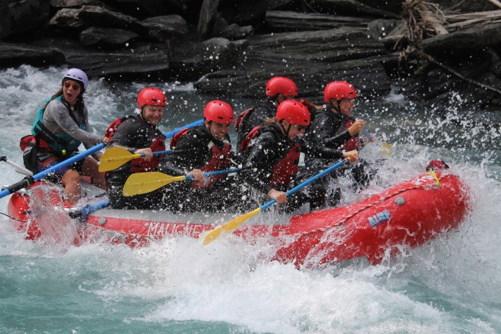Group of people in red helmets rafting through white-water rapids, splashing water around.