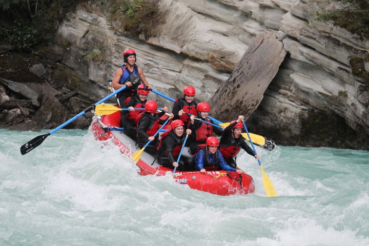 Group of people white-water rafting in a rocky river, wearing helmets and life jackets.