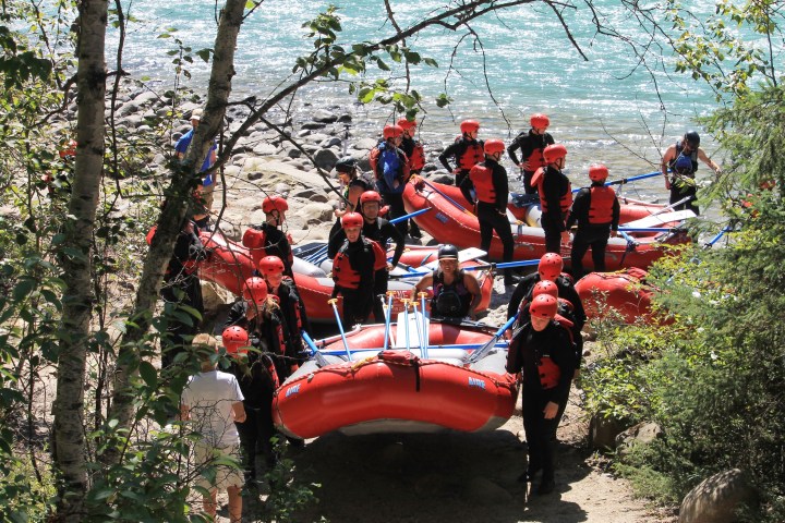 Group of people with red helmets and life vests holding rafts near a river.