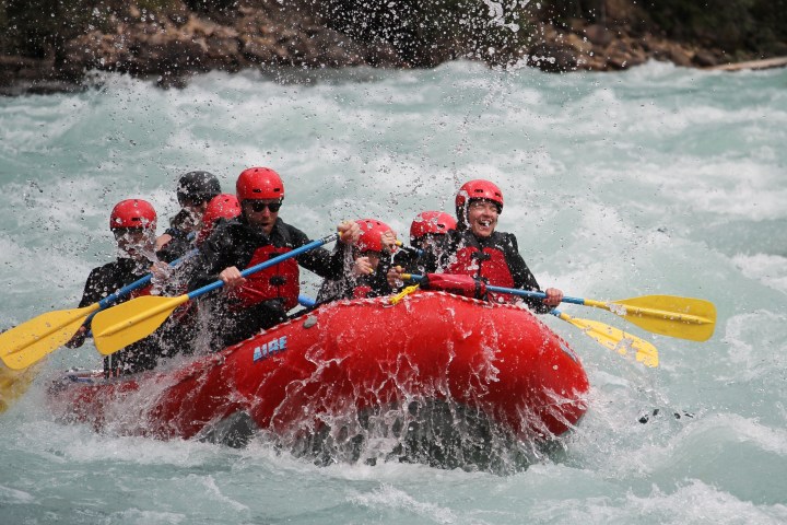 Group of people white-water rafting in a red inflatable boat with helmets and paddles.