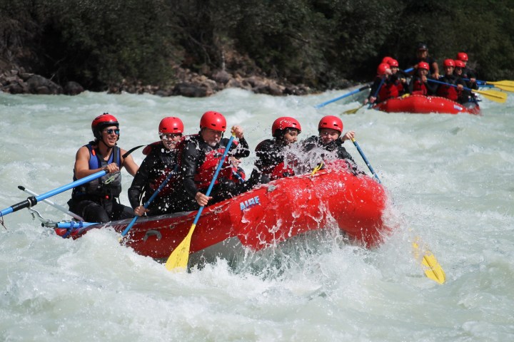 Group in red raft smiling and paddling in white-water rapids.