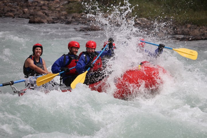 Group of people whitewater rafting, wearing helmets and life vests, with water splashing around them.