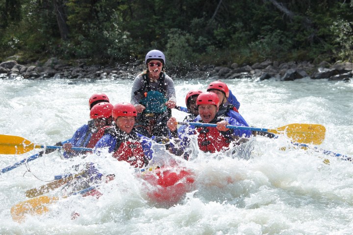 Group of people white-water rafting, wearing helmets and life jackets, surrounded by splashing water.