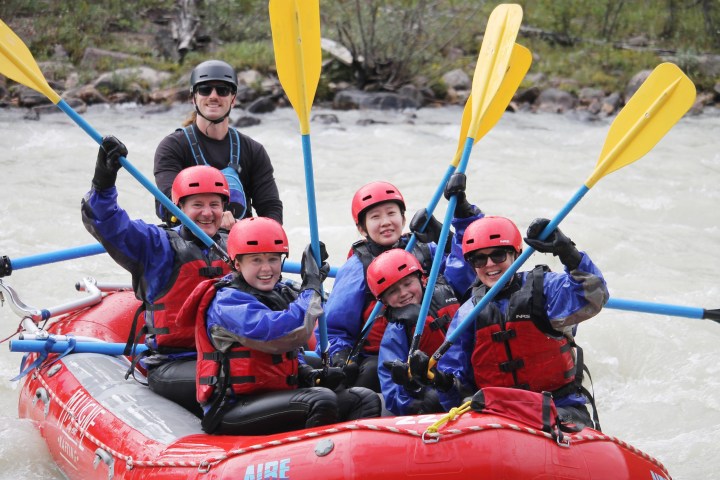 Group of six smiling people in helmets and life jackets rafting on a river, holding paddles up.
