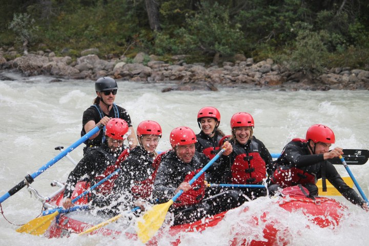 Group white water rafting on a river, all wearing helmets and life jackets, surrounded by splashing water.