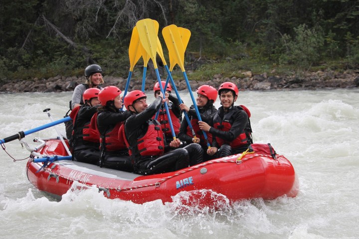 Group of six people rafting on a river with yellow paddles raised, wearing helmets and life vests.