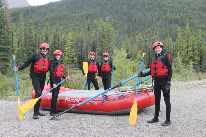 Five people in red vests and helmets with paddles near an inflatable raft in a forested area.