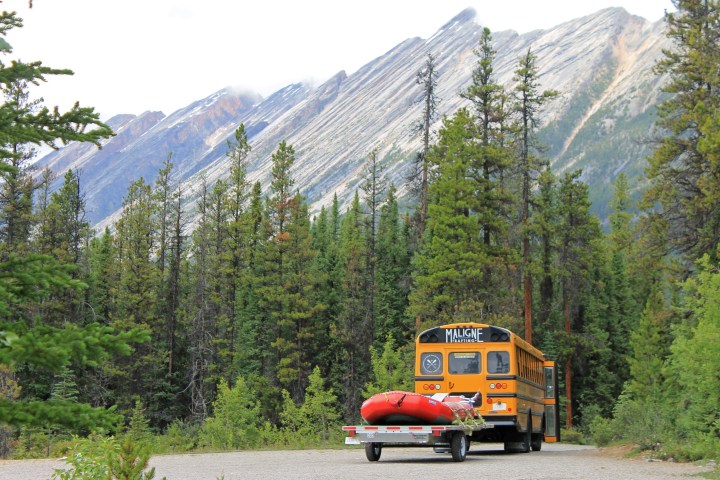 Bus with raft on trailer parked in front of tall trees and rocky mountains.