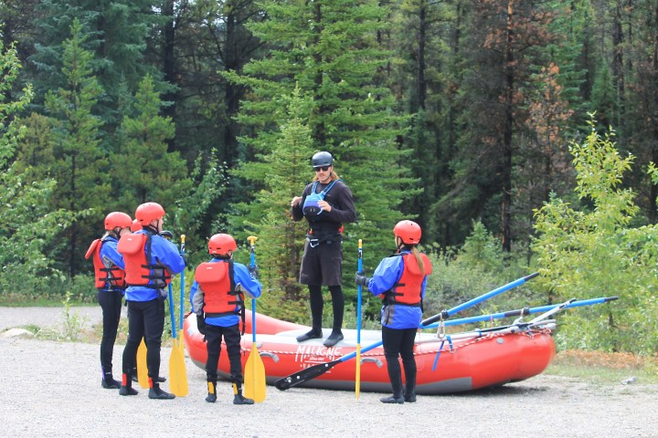 Group of people in helmets and life jackets near a red raft, surrounded by trees.