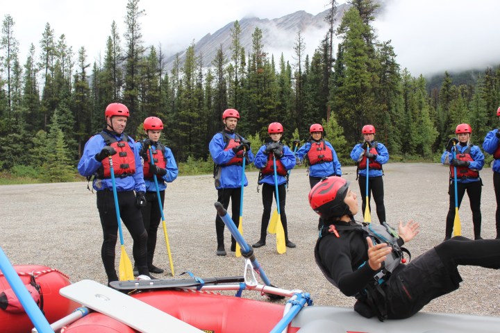 Group of people in rafting gear, listening to instructor in forested mountain area.