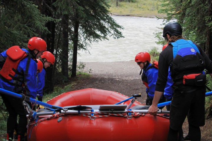 Four people in helmets and life vests prepare to launch a red raft toward a river.
